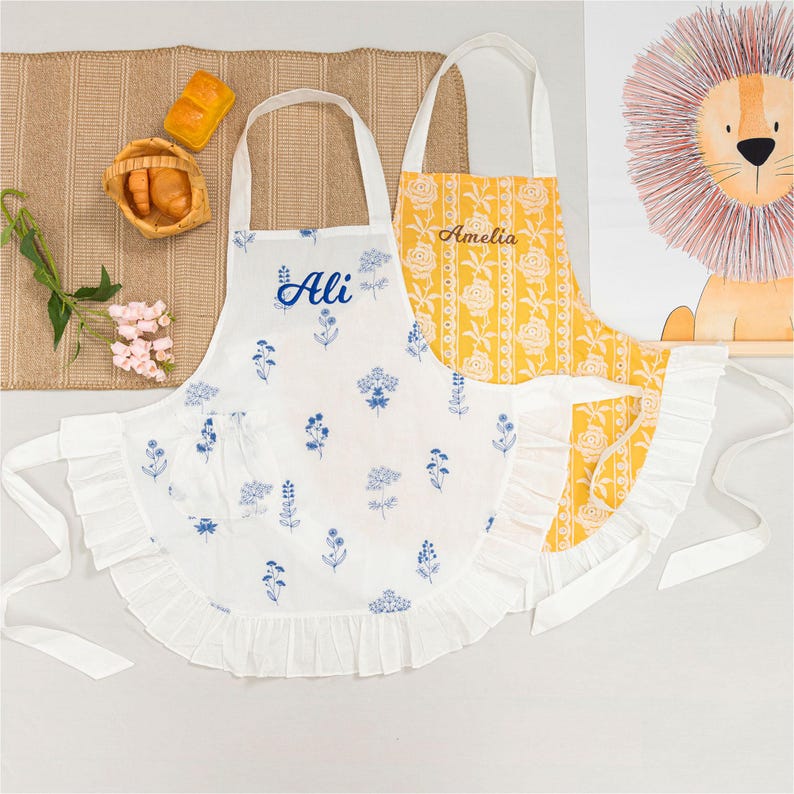 May include: Two aprons, one white and one yellow, are displayed. The white apron has blue floral patterns and the name "Ali" in blue script. The yellow apron features a floral pattern and the name "Amelia" in gold. Both aprons have white ties and ruffles.