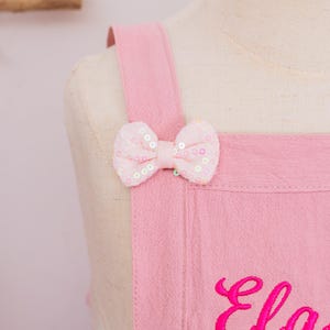 May include: Close-up of a pink apron with a bow detail. The bow is made of pink fabric and adorned with sequins. The apron has shoulder straps and an embroidered name in a cursive font.