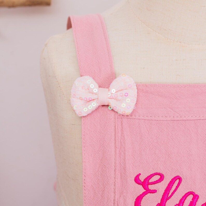 May include: Close-up of a pink apron with a bow detail. The bow is made of pink fabric and adorned with sequins. The apron has shoulder straps and an embroidered name in a cursive font.