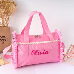May include: Pink duffel bag with a shoulder strap and handles. The bag features a white lace trim and ruffled edges. The name "Olivia" is embroidered in a matching pink thread. A small star-shaped charm adorns the zipper.