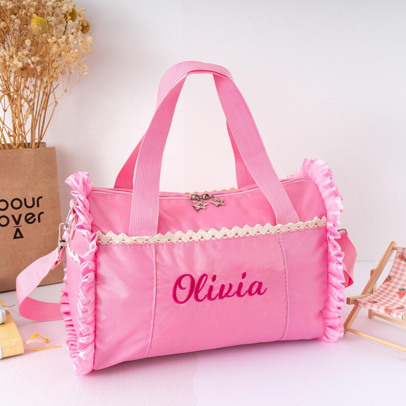 May include: Pink duffel bag with a shoulder strap and handles. The bag features a white lace trim and ruffled edges. The name "Olivia" is embroidered in a matching pink thread. A small star-shaped charm adorns the zipper.