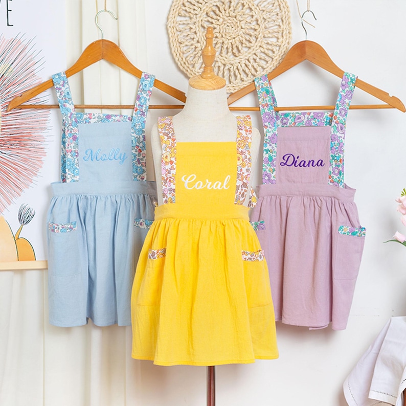May include: Three children's aprons in pastel colors: light blue, yellow, and lavender. Each apron has floral patterned straps and pocket accents. The names "Molly", "Coral", and "Diana" are embroidered on the aprons.