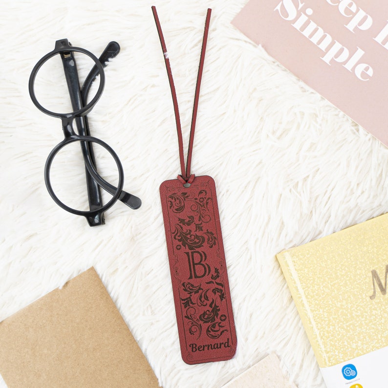 May include: A brown leather bookmark with a decorative floral design and the letter "B" engraved in the center. The bookmark also has the name "Bernard" engraved at the bottom.