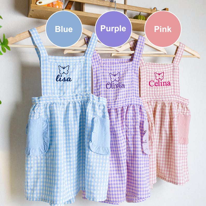 May include: Three gingham aprons with butterfly embroidery and personalized names. The aprons are blue, purple, and pink. The names are Lisa, Olivia, and Celina.