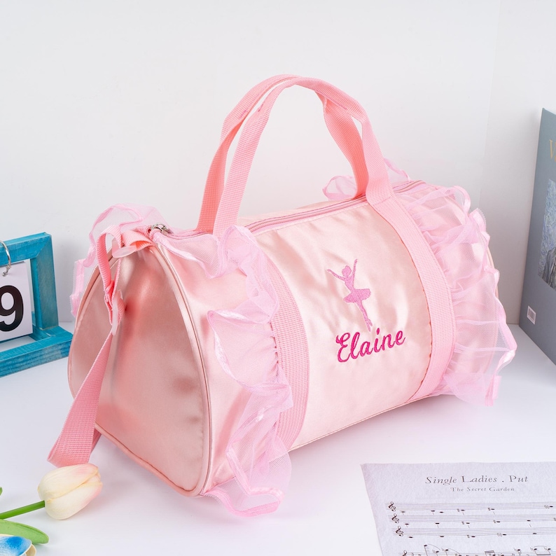 May include: Pink satin dance bag with a pink tulle ruffle trim. The bag has a pink ballerina silhouette and the name "Elaine" embroidered on the front.