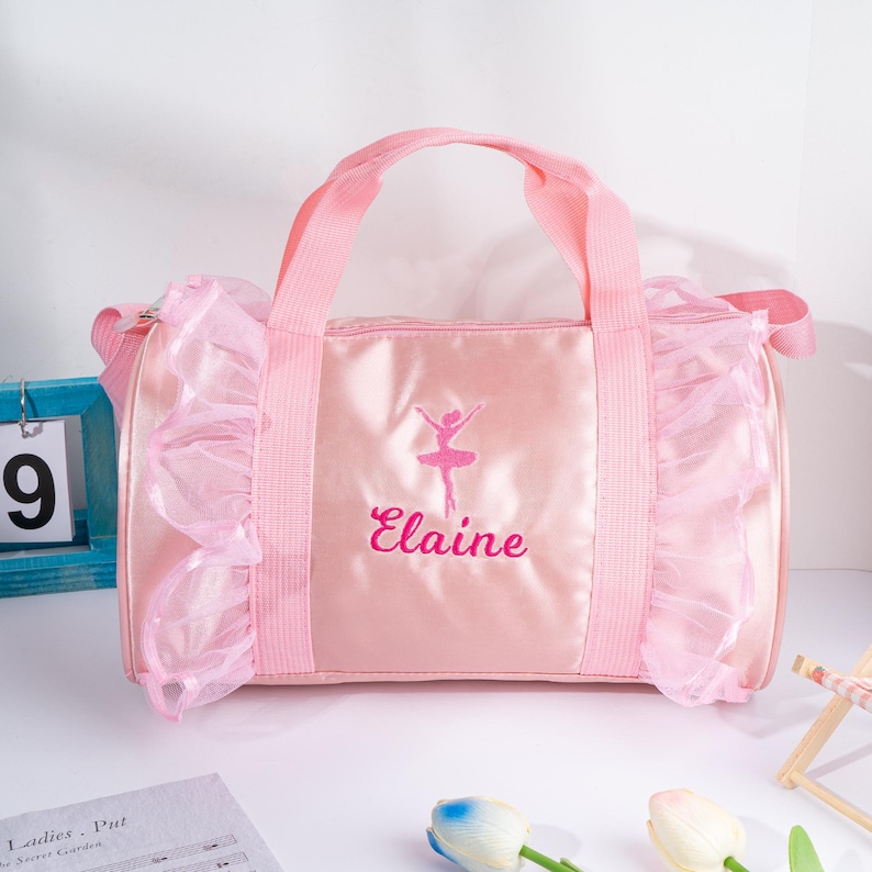 May include: Pink satin dance bag with a ballerina silhouette and the name "Elaine" embroidered on the front. The bag has a ruffled trim and a top handle.