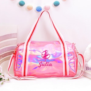 May include: Pink duffel bag with a holographic finish, featuring a ballerina graphic and the name "Julia" embroidered in cursive. The bag has red and white striped handles and a detachable shoulder strap.