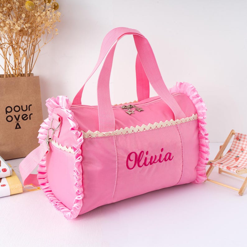 May include: Pink duffel bag with a top zipper, handles, and a shoulder strap. The bag has a decorative lace trim and ruffled edges. The name "Olivia" is embroidered on the front. A small star charm hangs from the zipper.