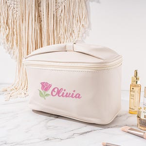 May include: A white faux leather cosmetic bag with a zipper closure. The bag has a pink rose and the name "Olivia" embroidered on the front.