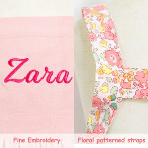 May include: A light pink item with the name "Zara" embroidered in a bright pink script. The item has floral patterned straps with pink, yellow, and green flowers on a white background. The text "Fine Embroidery" and "Floral patterned straps" are at the bottom.