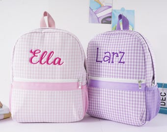 May include: Two backpacks with a gingham pattern. The left backpack is light pink and white with the name Ella embroidered in pink. The right backpack is light purple and white with the name Larz embroidered in purple. Both have a top handle.