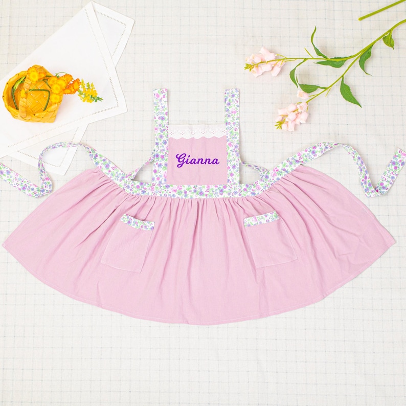 以下が含まれることがあります： ピンク色のエプロン。前面には「Gianna」の名前が刺繍され、花柄のアクセントが施されています。フリルスカート、2つのポケット、花柄のストラップとトリムが特徴です。キッチンやクラフト用途に適しています。