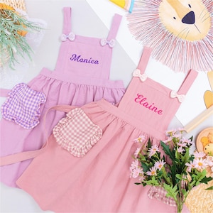 May include: Two pink aprons with bow accents and embroidered names, "Monica" and "Elaine". Each apron has a ruffled skirt and a gingham pocket. A bouquet of pink flowers and a lion illustration are in the background.