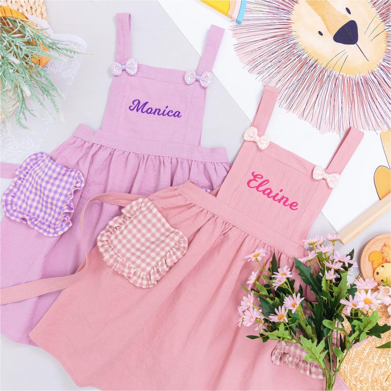 May include: Two pink aprons with bow accents and embroidered names, "Monica" and "Elaine". Each apron has a ruffled skirt and a gingham pocket. A bouquet of pink flowers and a lion illustration are in the background.