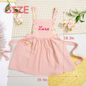 May include: A pink apron with floral patterned straps and pocket details. The apron has the name "Zara" embroidered on the front. The apron measures 39.4 inches wide and 18.3 inches long.