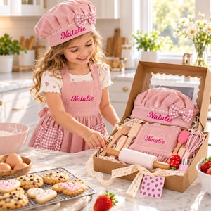 May include: A pink children's apron and chef hat set with white gingham accents, personalized with the name Natalie in pink embroidery. The set is presented in a cardboard gift box containing a wooden rolling pin, spatula, and whisk.