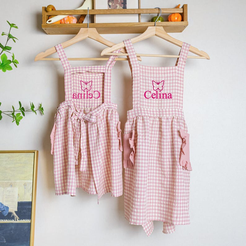 May include: Two pink and white gingham aprons with butterfly and name embroidery. The aprons have ruffled pockets and ties. The aprons are hanging on wooden hangers.