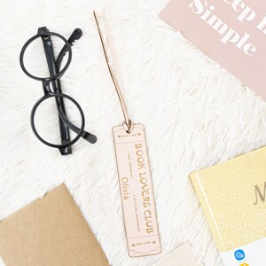 May include: A light brown wooden bookmark shaped like a ticket with the text "BOOK LOVERS CLUB" and "Olivia" printed on it. The bookmark has a brown leather strap.
