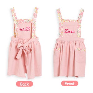 May include: Pink children's apron with floral patterned straps and pocket trim. The apron has the name "Zara" embroidered on the front and "Avia" on the back. A bow detail is tied at the waist. The apron is shown against a white background.
