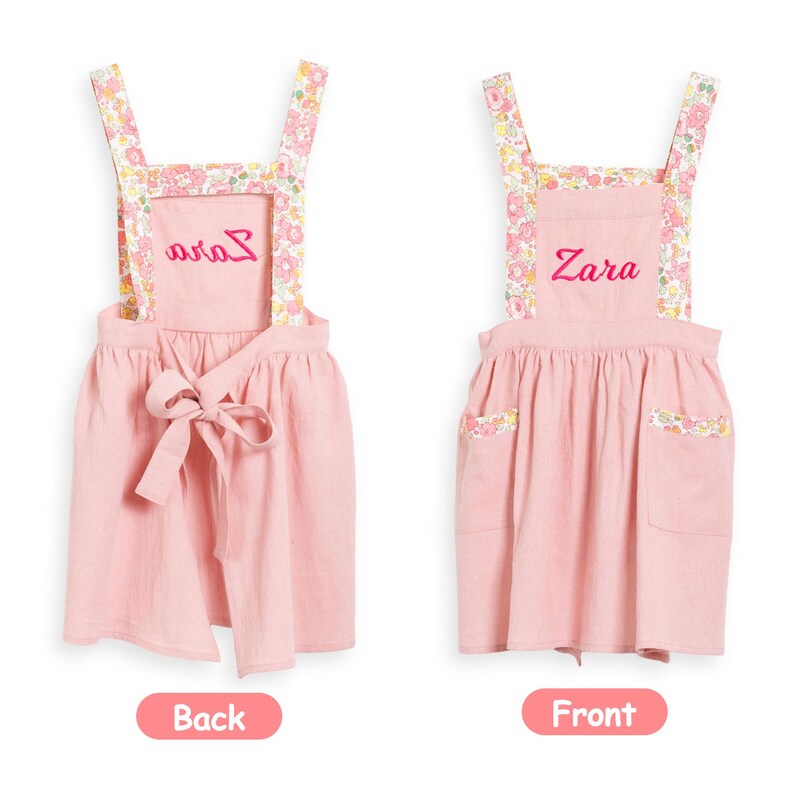 May include: Pink children's apron with floral patterned straps and pocket trim. The apron has the name "Zara" embroidered on the front and "Avia" on the back. A bow detail is tied at the waist. The apron is shown against a white background.