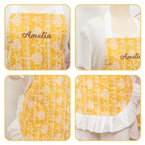 May include: A yellow apron with a floral pattern and the name "Amelia" embroidered in a cursive font. The apron has white trim and a ruffled bottom edge. The apron is displayed on a mannequin.