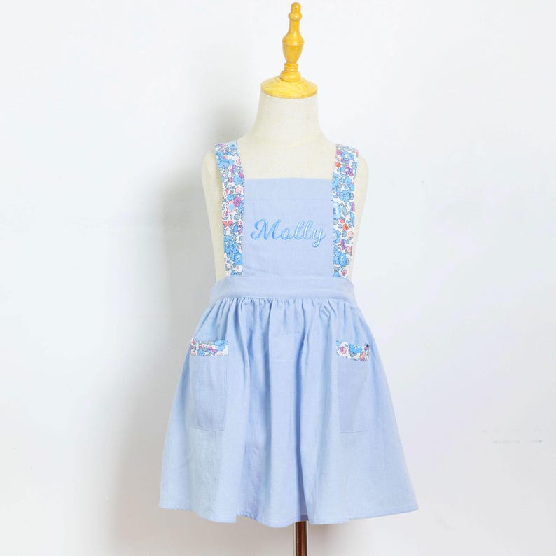 May include: A light blue apron with floral patterned straps and pocket accents. The name "Molly" is embroidered on the front. The apron is displayed on a mannequin.