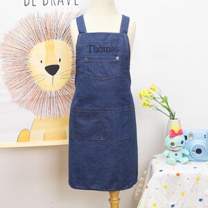 Peut inclure: Tablier en jean bleu avec le nom "Thomas" brod&eacute; sur la poche de poitrine. Le tablier a deux grandes poches avant et des bretelles crois&eacute;es. Le tablier est pr&eacute;sent&eacute; sur un mannequin. L'arri&egrave;re-plan comprend une illustration de lion et une petite table avec un jouet et des fleurs.