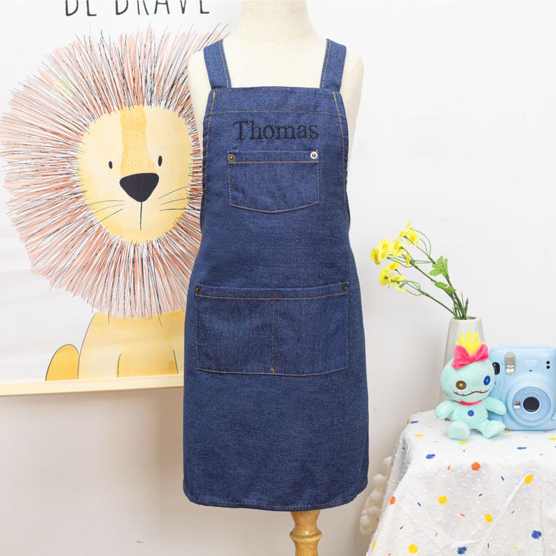 Peut inclure: Tablier en jean bleu avec le nom "Thomas" brod&eacute; sur la poche de poitrine. Le tablier a deux grandes poches avant et des bretelles crois&eacute;es. Le tablier est pr&eacute;sent&eacute; sur un mannequin. L'arri&egrave;re-plan comprend une illustration de lion et une petite table avec un jouet et des fleurs.