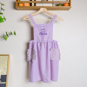 May include: A purple and white gingham apron with a butterfly and the name "Olivia" embroidered on the front. The apron has two ruffled pockets.