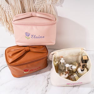 May include: Three personalized cosmetic bags with a bow detail. The bags are pink, brown, and white. The pink bag has the name "Elaine" embroidered on it, the brown bag has the name "Bonnie" embroidered on it, and the white bag has the name "Olivia" embroidered on it.