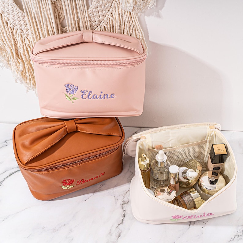 May include: Three personalized cosmetic bags with a bow detail. The bags are pink, brown, and white. The pink bag has the name "Elaine" embroidered on it, the brown bag has the name "Bonnie" embroidered on it, and the white bag has the name "Olivia" embroidered on it.