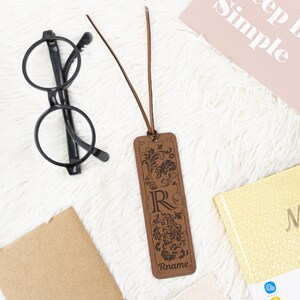 May include: A brown leather bookmark with a decorative floral design and the letter "R" engraved in the center. The bookmark has a brown leather tassel and the name "Rname" is engraved at the bottom.