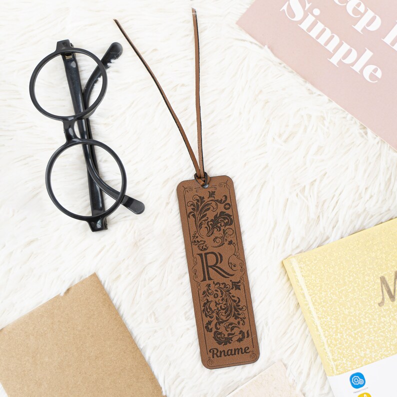 May include: A brown leather bookmark with a decorative floral design and the letter "R" engraved in the center. The bookmark has a brown leather tassel and the name "Rname" is engraved at the bottom.