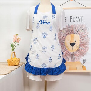 May include: White apron with blue floral print and the name "Vera" in script. The apron has a blue ruffle along the bottom and blue straps. A pocket is on the front left side. The apron is displayed on a mannequin.