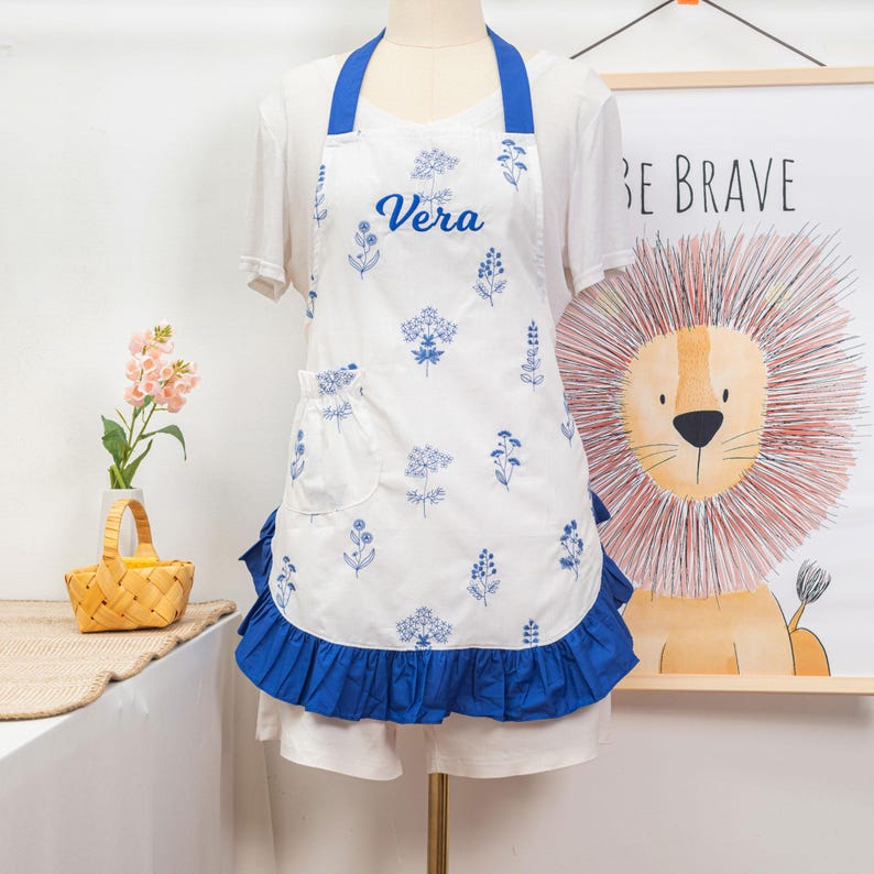 May include: White apron with blue floral print and the name "Vera" in script. The apron has a blue ruffle along the bottom and blue straps. A pocket is on the front left side. The apron is displayed on a mannequin.