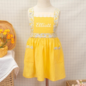 以下が含まれることがあります： 白で「Elliott」と刺繍された明るい黄色のエプロン。ストラップ、ウエスト、ポケットに花柄のアクセントがあります。白いレースのトリムが上部を飾っています。マネキンにエプロンが表示されています。