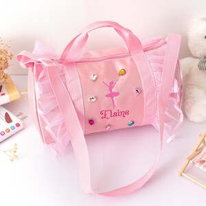 Peut inclure: Sac de sport rose avec un motif de ballerine et le nom "Elaine". Le sac a une poign&eacute;e sup&eacute;rieure, une bandouli&egrave;re et est orn&eacute; de strass et de volants. Un joli sac pour une jeune danseuse.