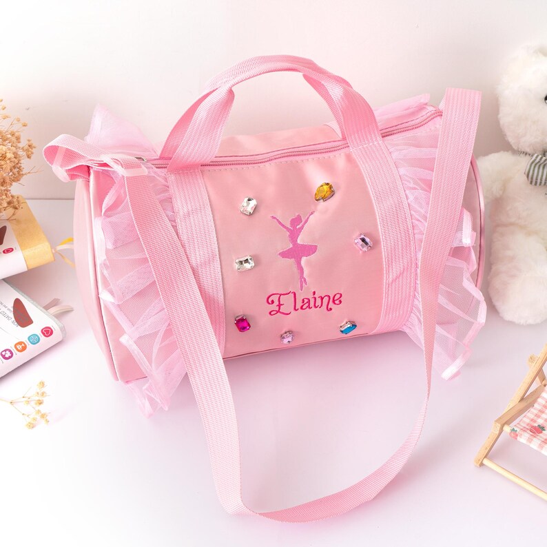 Peut inclure: Sac de sport rose avec un motif de ballerine et le nom "Elaine". Le sac a une poign&eacute;e sup&eacute;rieure, une bandouli&egrave;re et est orn&eacute; de strass et de volants. Un joli sac pour une jeune danseuse.