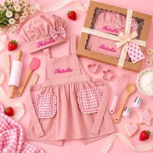 May include: A pink children's apron and chef hat set personalized with the name Natalie in pink embroidery. The set includes a matching pink gingham pocket apron, a pink whisk, a wooden rolling pin, and a gift box with a strawberry tag.