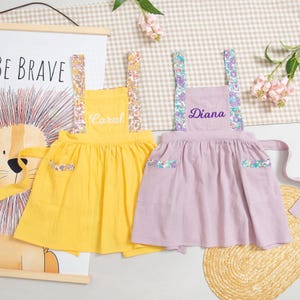 May include: Two children's aprons, one yellow and one lavender, with floral patterned straps and pocket accents. The yellow apron has the name "Coral" embroidered on it, while the lavender apron has "Diana". The aprons are displayed on a light-colored surface.