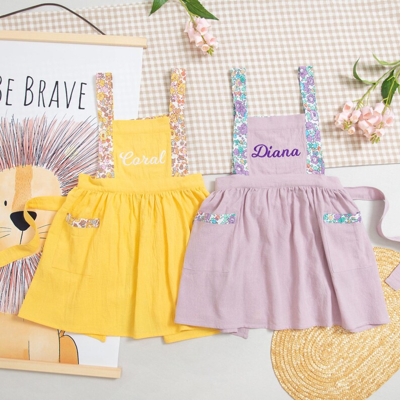 May include: Two children's aprons, one yellow and one lavender, with floral patterned straps and pocket accents. The yellow apron has the name "Coral" embroidered on it, while the lavender apron has "Diana". The aprons are displayed on a light-colored surface.