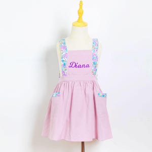 May include: A light pink apron with floral patterned straps and pocket accents. The name "Diana" is embroidered in purple script across the chest. The apron is displayed on a mannequin.