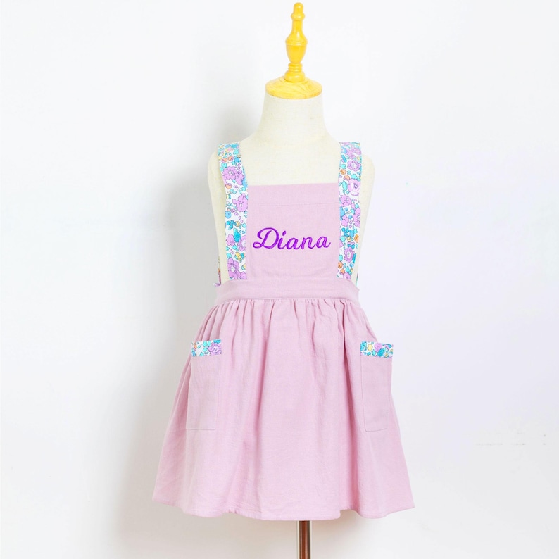 May include: A light pink apron with floral patterned straps and pocket accents. The name "Diana" is embroidered in purple script across the chest. The apron is displayed on a mannequin.