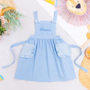 May include: A light blue apron with the name "Bonnie" embroidered on the chest. The apron has two pockets with a white and blue gingham pattern and ruffled edges. Small bows adorn the straps.