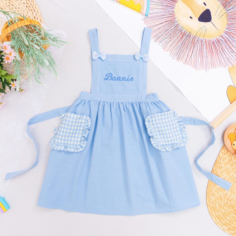 May include: A light blue apron with the name "Bonnie" embroidered on the chest. The apron has two pockets with a white and blue gingham pattern and ruffled edges. Small bows adorn the straps.