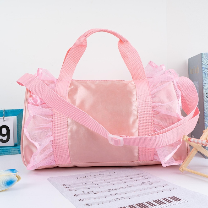 May include: Pink satin and tulle dance bag with a single strap and a ruffled trim. The bag has a zipper closure and a small pocket on the front.