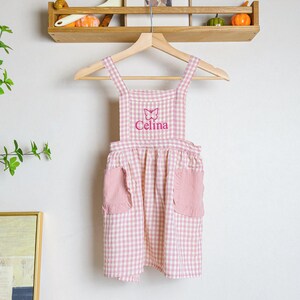 May include: Pink and white gingham apron with a butterfly and the name "Celina" embroidered on the front. The apron has two pockets on the front.