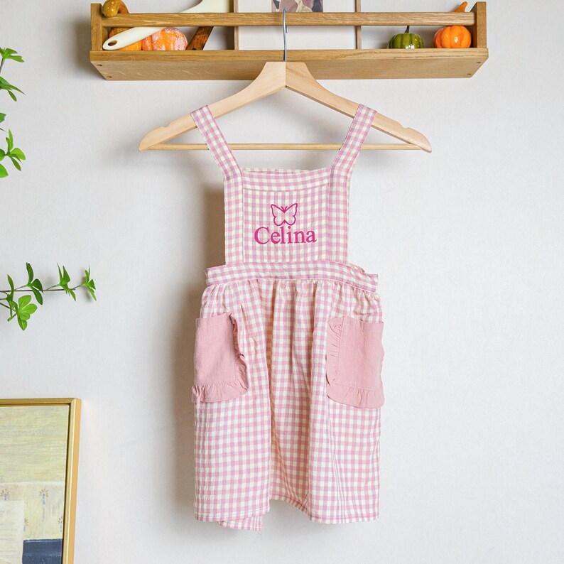 May include: Pink and white gingham apron with a butterfly and the name "Celina" embroidered on the front. The apron has two pockets on the front.