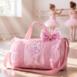 May include: A pink duffel bag featuring a sequined ballerina appliqué, the name Sandra embroidered in script, and a large glittery pink bow. The bag is accented with ruffled tulle trim on the sides and includes a matching shoulder strap.