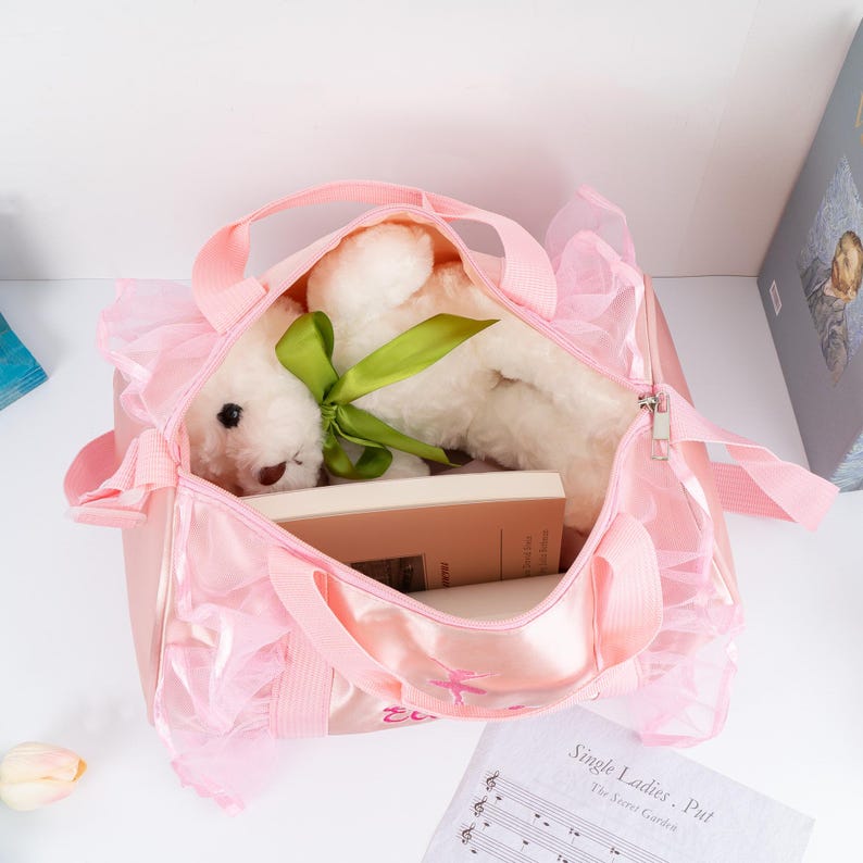 May include: Pink ballerina-themed duffel bag with a white teddy bear inside. The bag has a ruffled trim and a zipper closure. The bag is embroidered with a ballerina and the words "En Pointe".